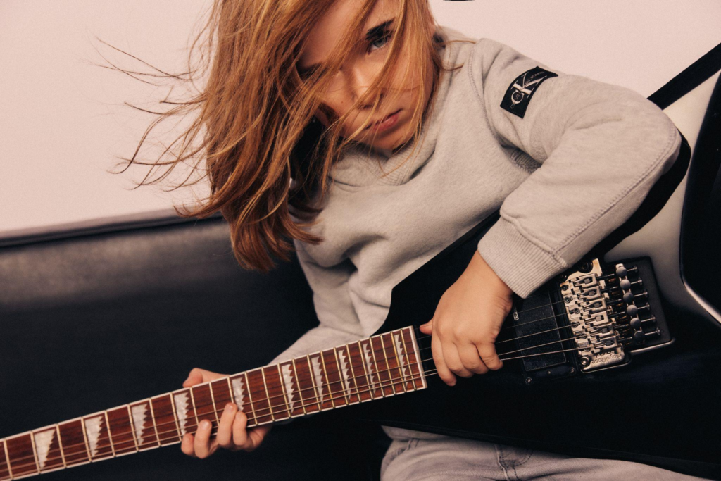 Chłopiec z długimi włosami grający na gitarze w szarej bluzie Calvin Klein