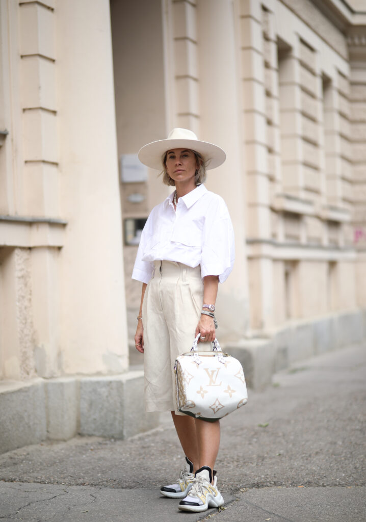 Kobieta w beżowych spodniach i luźnej koszuli w kapeluszu i białą torebką