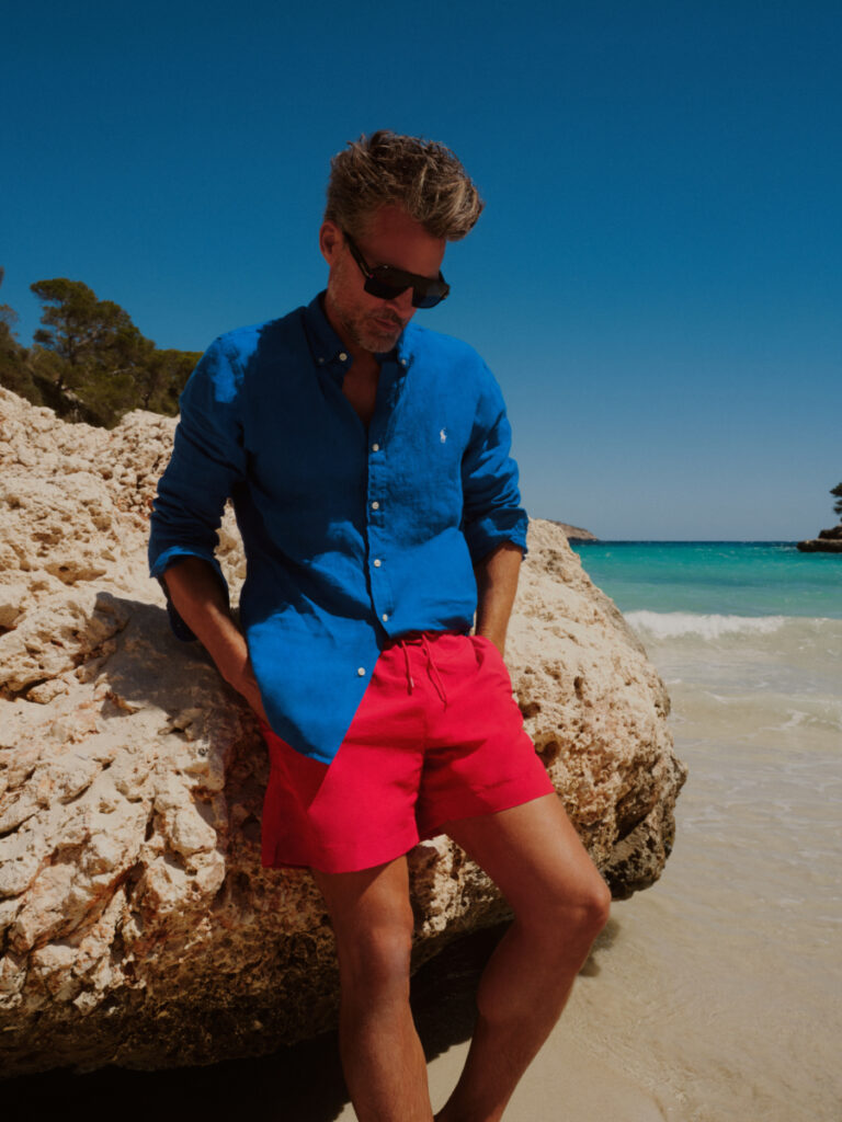 mężczyzna na plaży w czerwonych szortach kąpielowych, niebieskiej koszuli i okularach przeciwsłonecznych